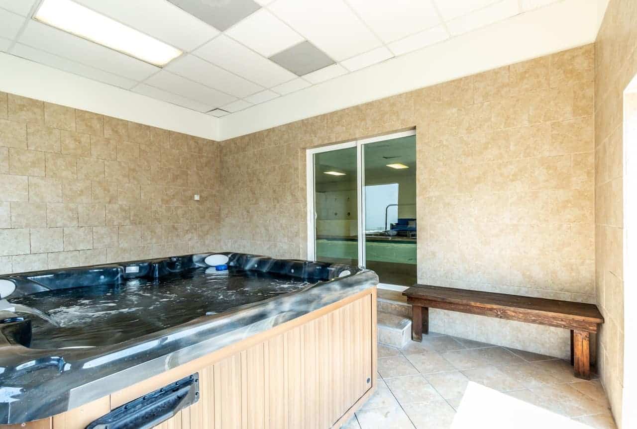 Indoor room with a large black hot tub, tan tiled walls and floor, a wooden bench against the wall, and glass doors leading to another room with a pool—experience comfort at Alpine Chalet Apartments Rexburg.