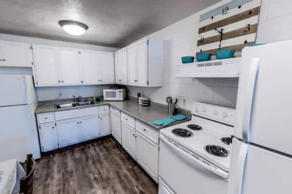 Bright kitchen at Alpine Chalet Apartments Rexburg with white cabinets, appliances, and countertops. Features a microwave, teal kitchenware, and a large wall clock above the stove. Wood-patterned floors and ample lighting complete the space.