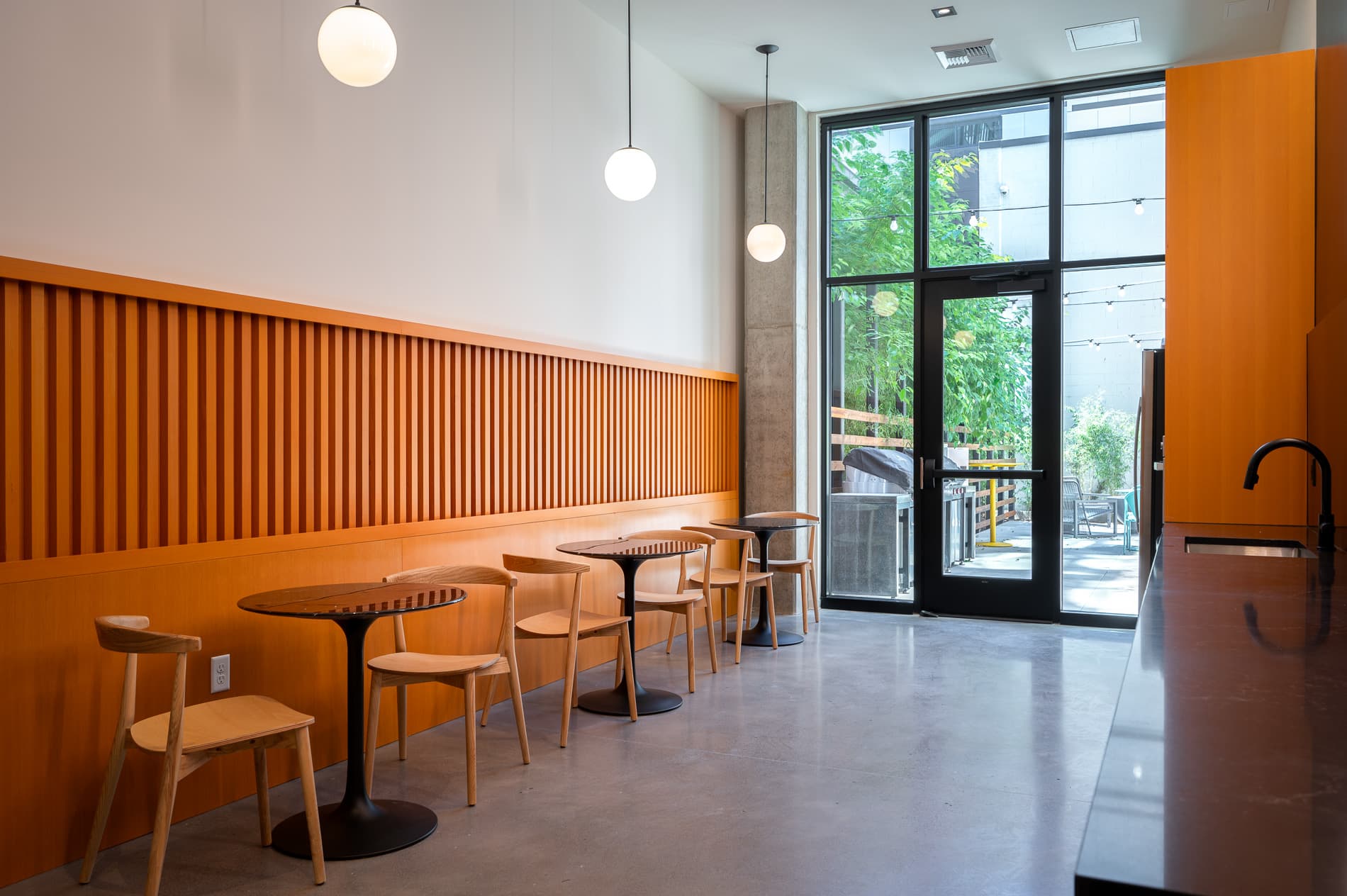 A modern, minimalist cafe interior at MERX Slabtown features wooden tables and chairs, orange paneling along the wall, hanging globe lights, and large glass doors opening to a lush outdoor patio area.