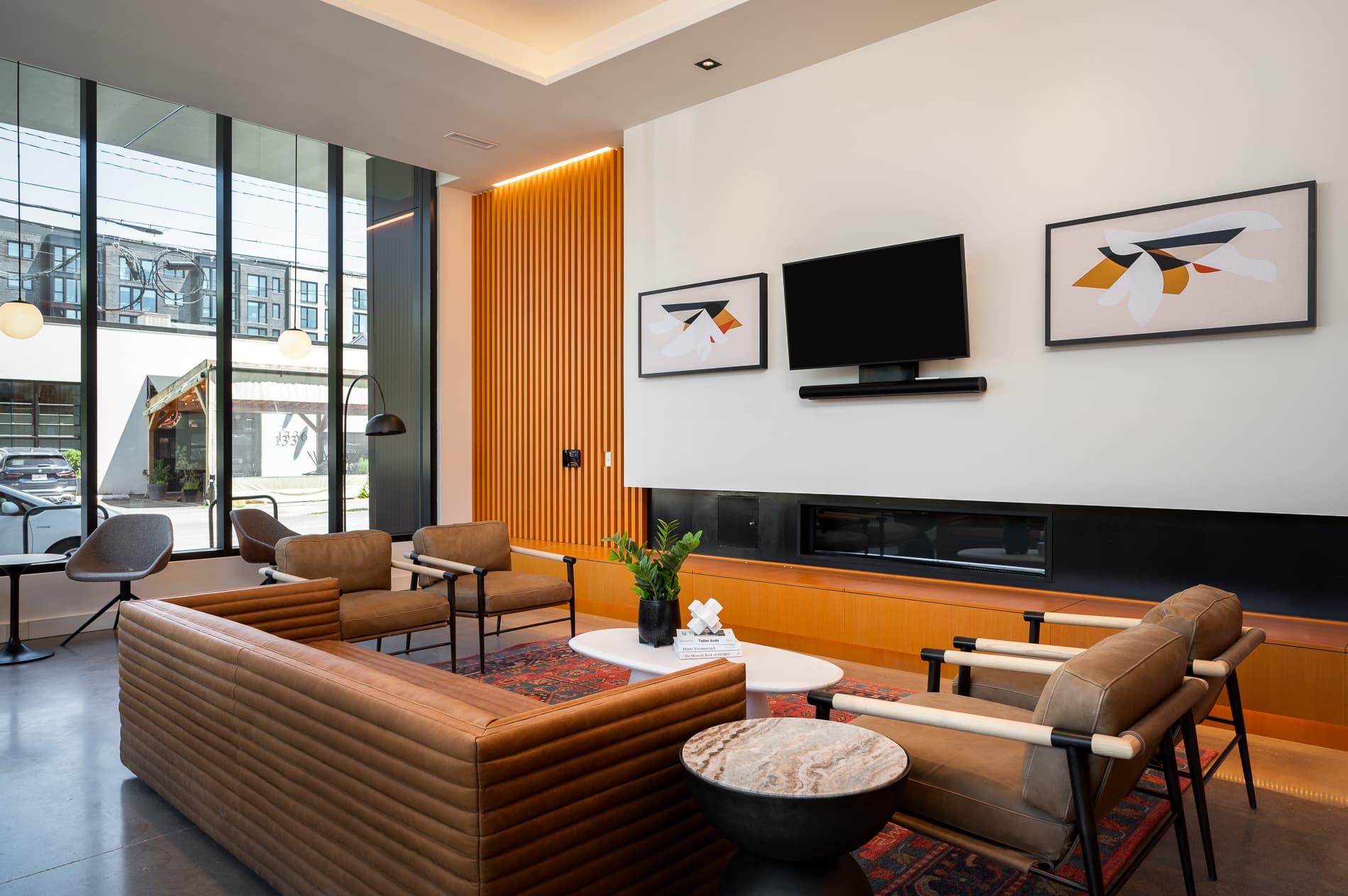 A modern Slabtown lobby with brown leather sofas, armchairs, a round coffee table, and a wall-mounted TV between two abstract art pieces. Large windows let in natural light, and wood slats accent one wall.