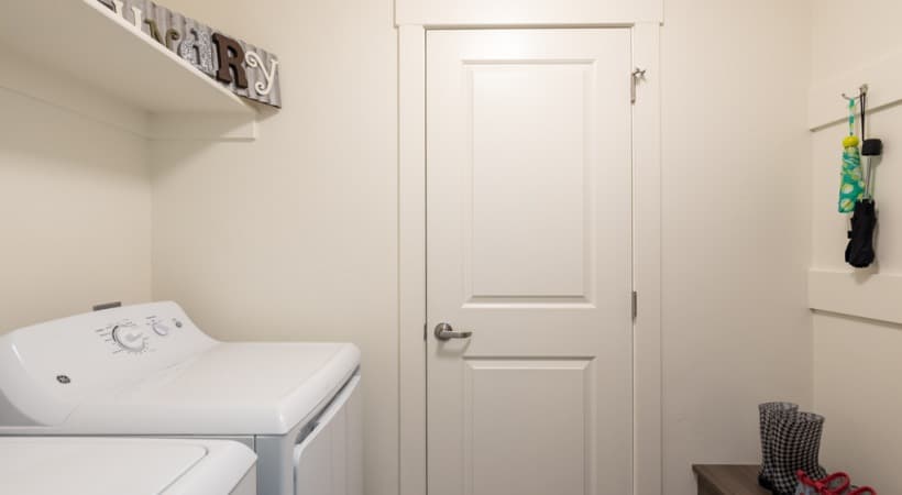 A small laundry room at Arboretum at Barber Station Boise features a white washing machine and dryer, a closed white door, and a shelf with decorative items. Hooks on the wall hold gloves and a dog leash; boots are on the floor.