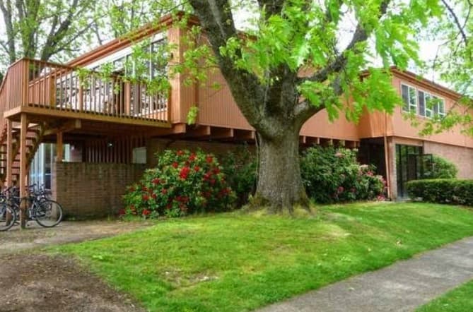 A two-story brown iHouse with large windows, a wooden deck and stairs, surrounded by green grass, trees, bushes, and red flowers in Corvallis, with two bicycles parked near the building.