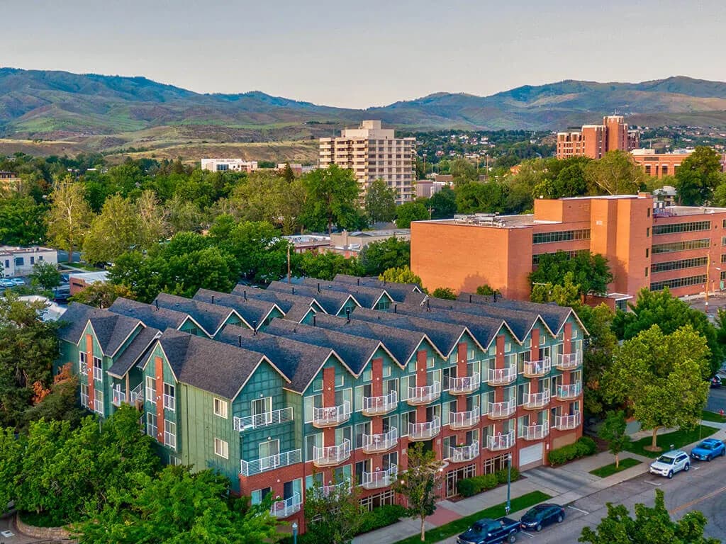 Aerial view of C W Moore Apartments Boise, a residential building with green siding and red accents, surrounded by trees and neighboring brick buildings, set against rolling green hills and more buildings beneath a clear sky.