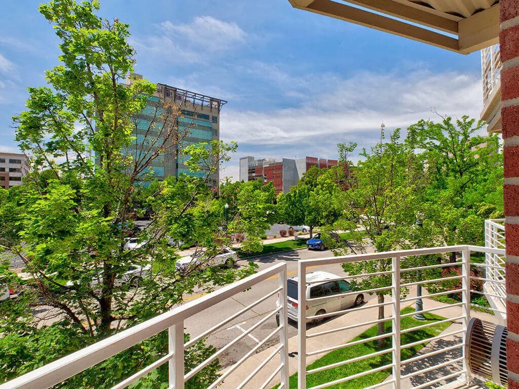 View from a balcony with a white railing at C W Moore Apartments Boise, overlooking a tree-lined street, parked cars, and office buildings under a blue sky with scattered clouds.