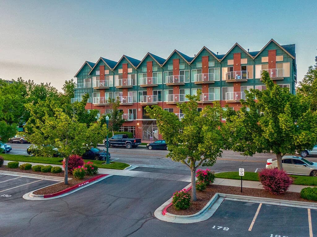 A modern, three-story apartment building—C W Moore Apartments Boise—with balconies and large windows faces a tree-lined street with parked cars. In the foreground, an empty parking lot with numbered spaces and landscaped areas is visible.