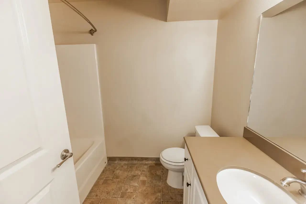A small bathroom with beige walls, tiled floor, a bathtub with a shower curtain rod, toilet, and a vanity with sink and large mirror. The clean, simple space offers practicality for residents of Campus Park Rexburg. Minimal decor adds to its charm.