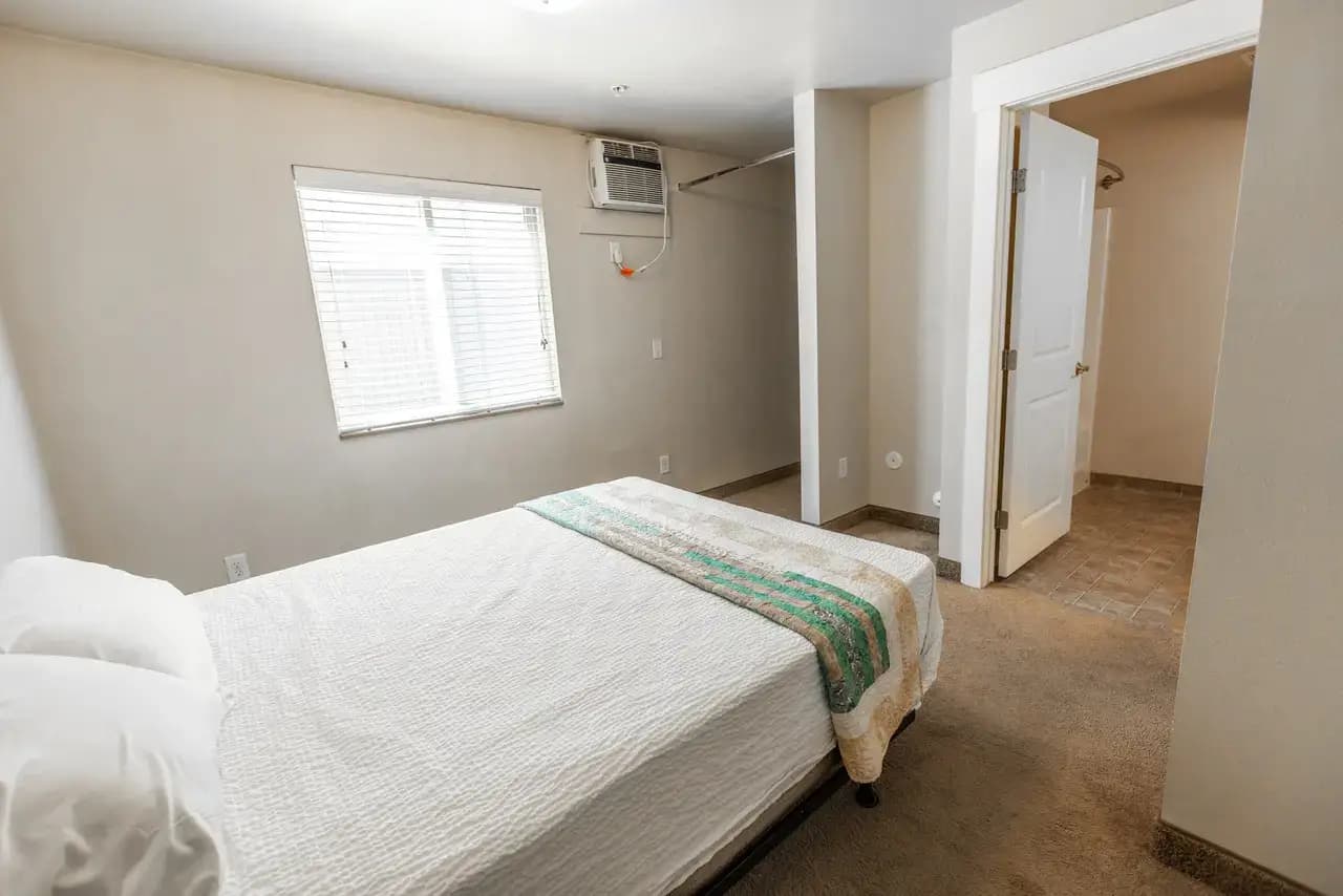 A simple bedroom at Campus Park Rexburg with a bed covered in white bedding, a colorful striped throw, a window with blinds and air conditioner above, closet space, beige carpet, and an open door leading to another room.