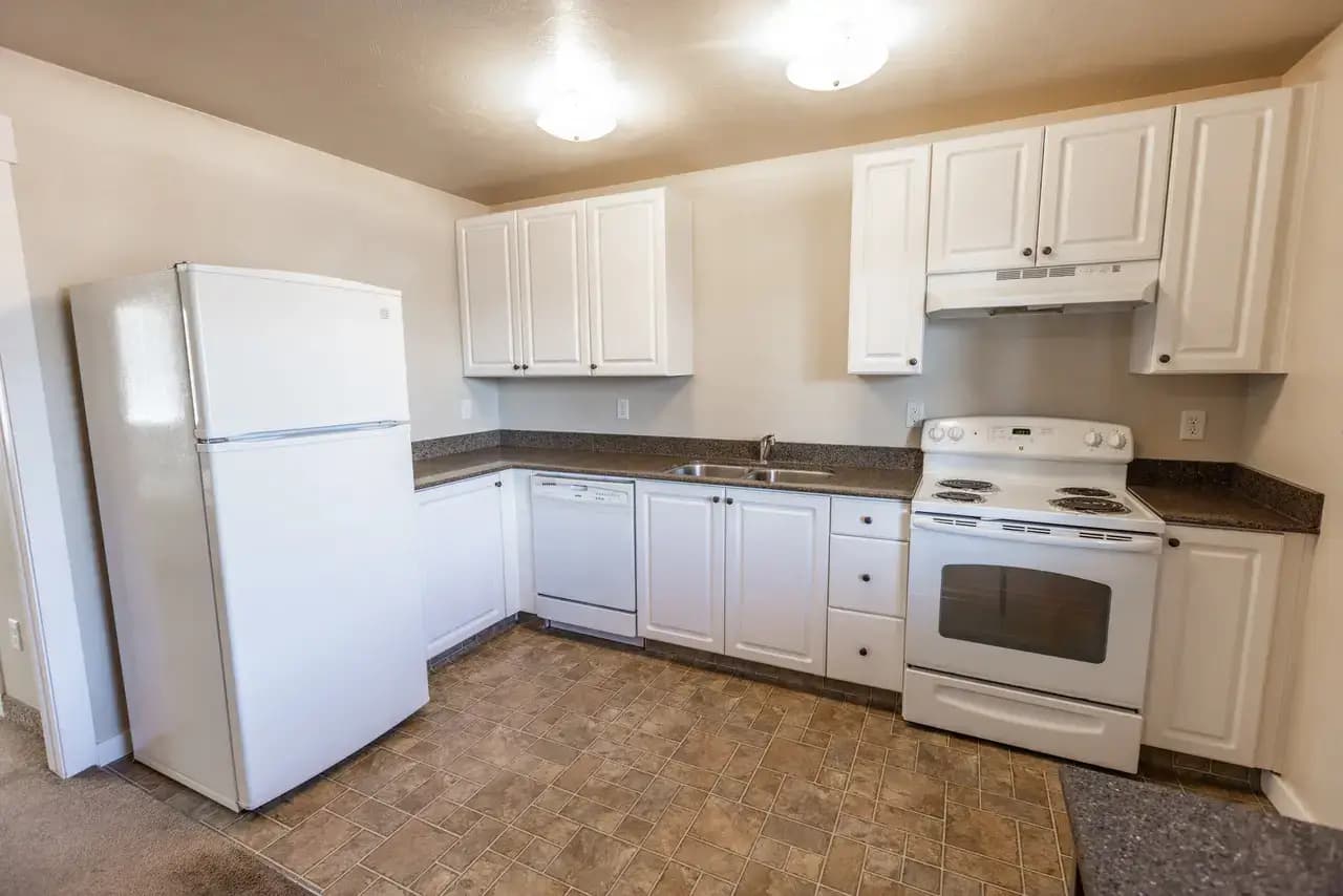 A clean, empty kitchen with white cabinets, a white refrigerator, dishwasher, electric stove with oven, range hood, and double sink—all set against beige walls and a brown-tiled floor—perfect for your Campus Park Rexburg home.