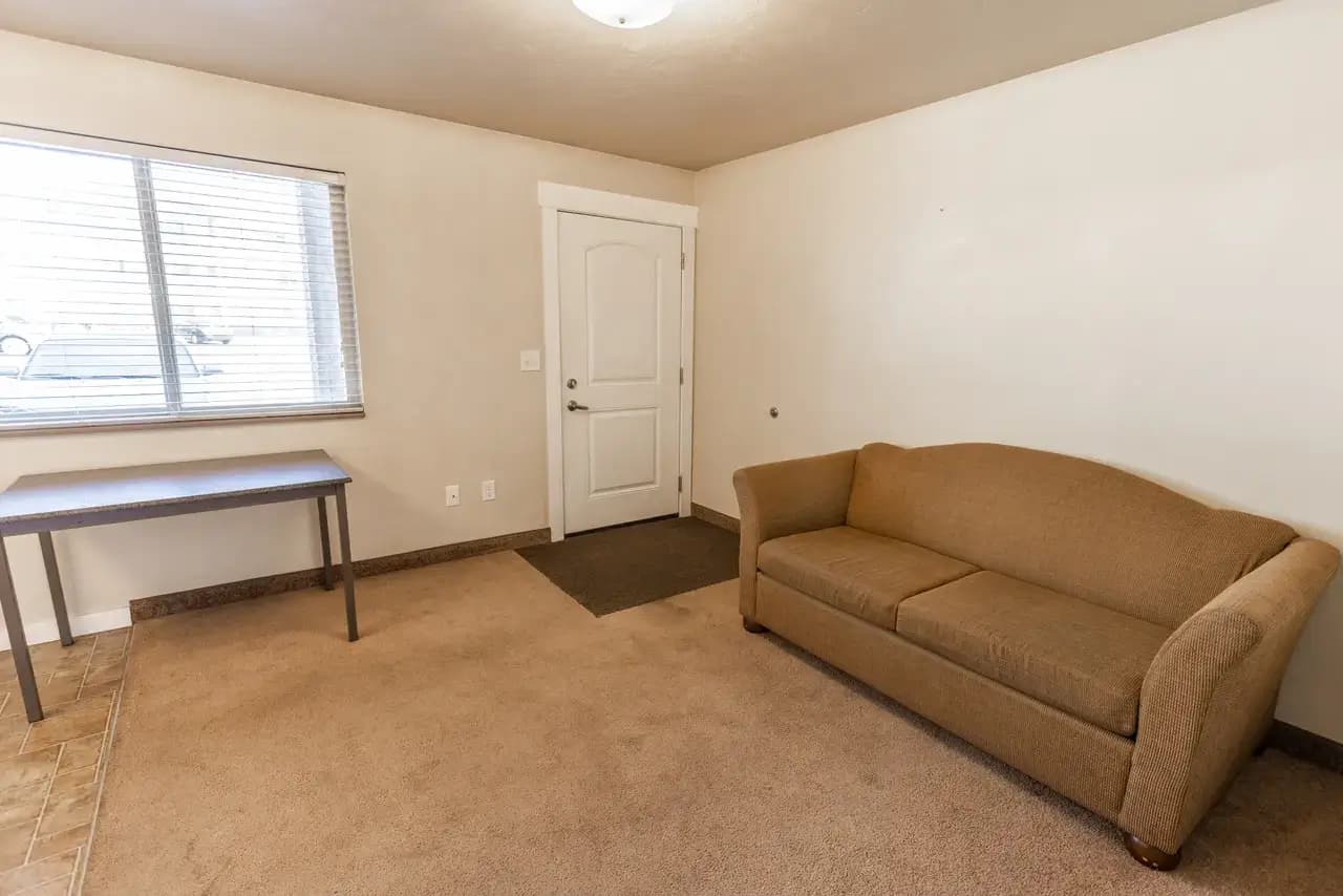 A simple room with beige carpet, a tan sofa, a small dark wood table by a window with blinds, and a white door on the far wall. The cream-colored walls and minimal furnishings evoke the calm of Campus Park Rexburg.