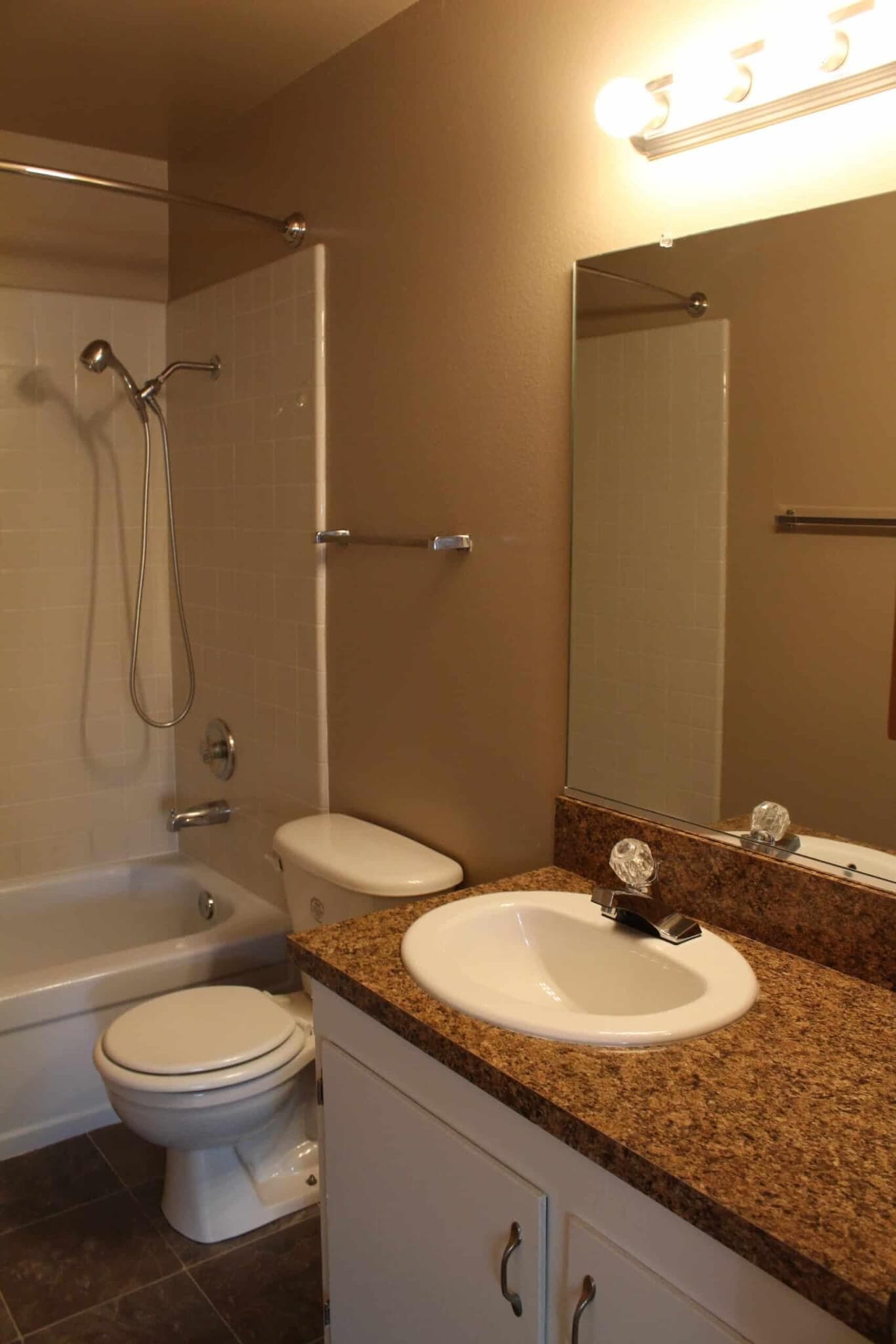 A bathroom at Campus Place Apartments Chico features a shower-tub combo, toilet, and a sink with brown granite countertop. A large mirror and three-bulb light fixture sit above the sink, with beige walls and dark tile flooring.