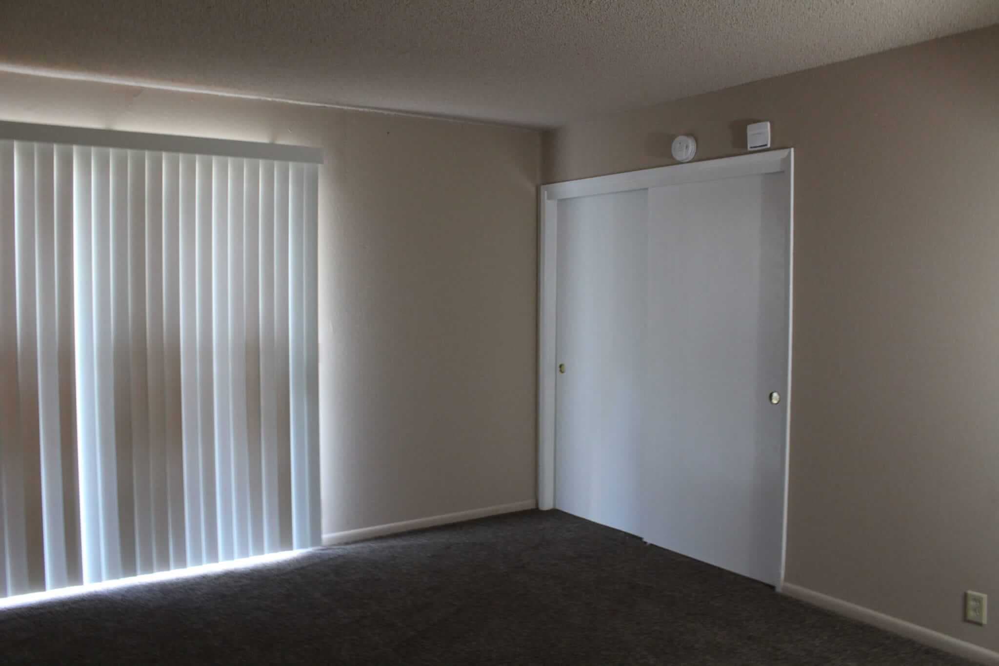 An empty room at Campus Place Apartments Chico features beige walls, gray carpet, a sliding glass door with vertical blinds on the left, and a white sliding closet door on the right.
