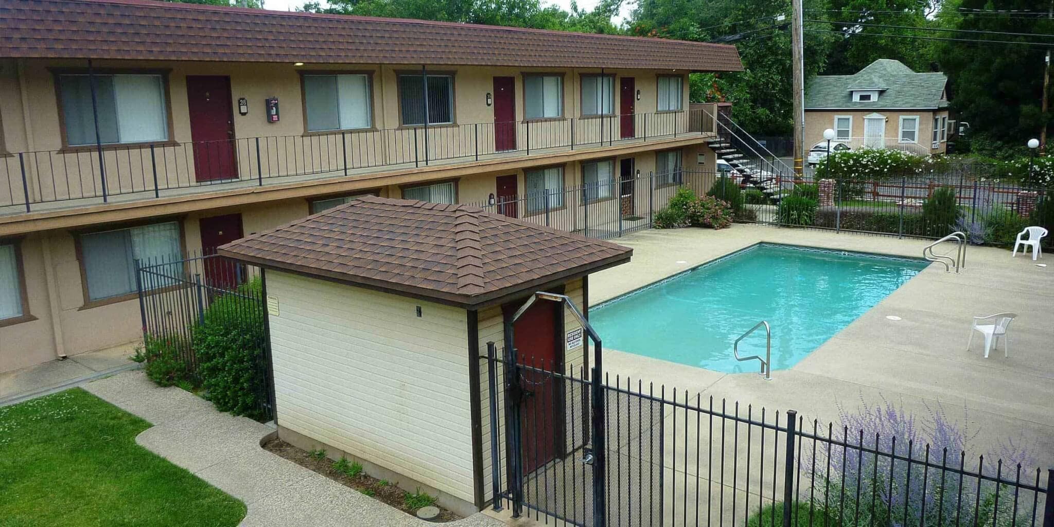 Campus Place Apartments Chico features a small two-story complex with red doors and outdoor walkways surrounding a fenced-in swimming pool. A few white chairs sit on the pool deck, with grass and trees visible in the background.