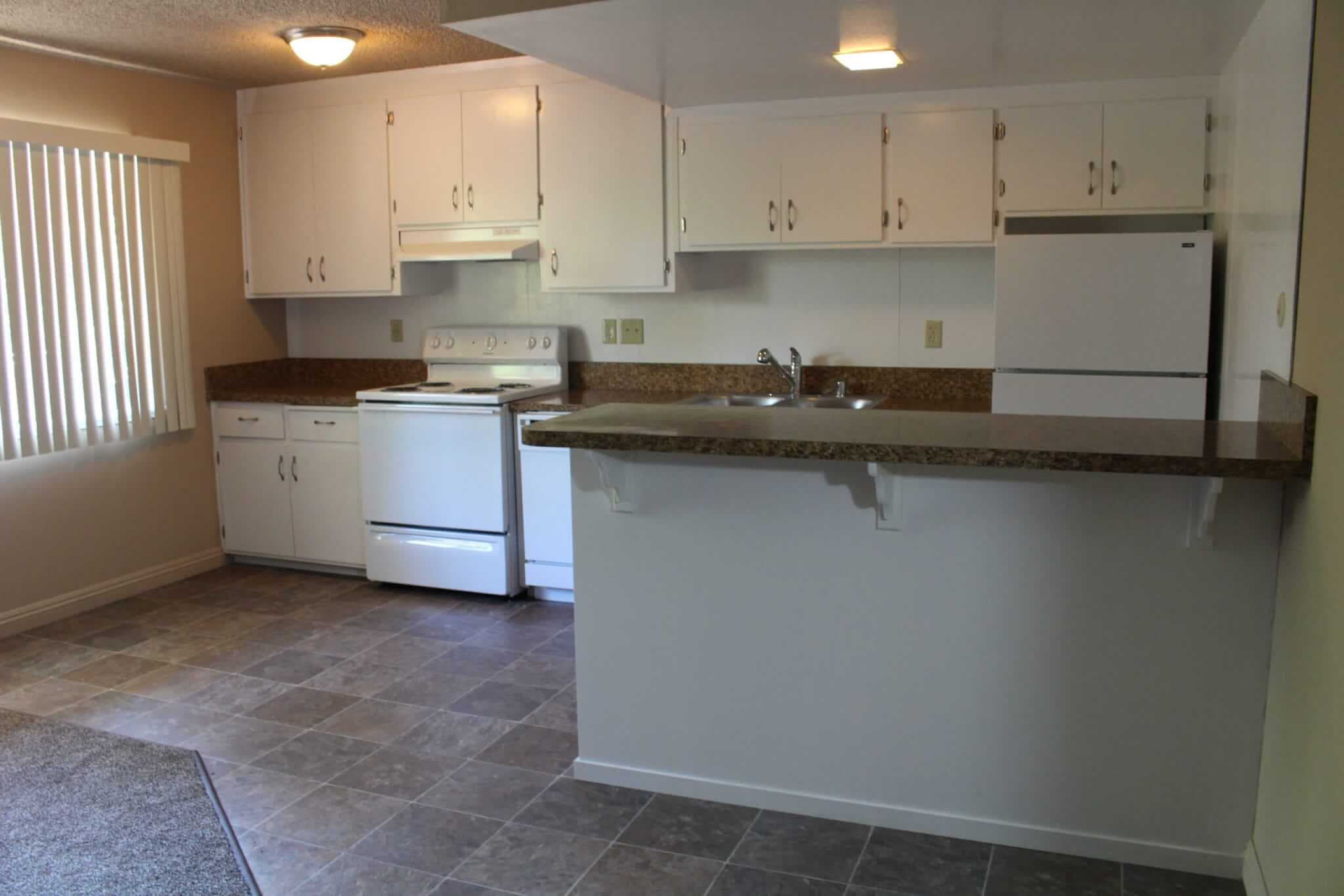 Apartment kitchen at Campus Place Apartments Chico features white cabinets, stove, fridge, and range hood. Brown countertops with a breakfast bar, tiled floor, vertical blinds on the window, neutral walls, and overhead lighting.