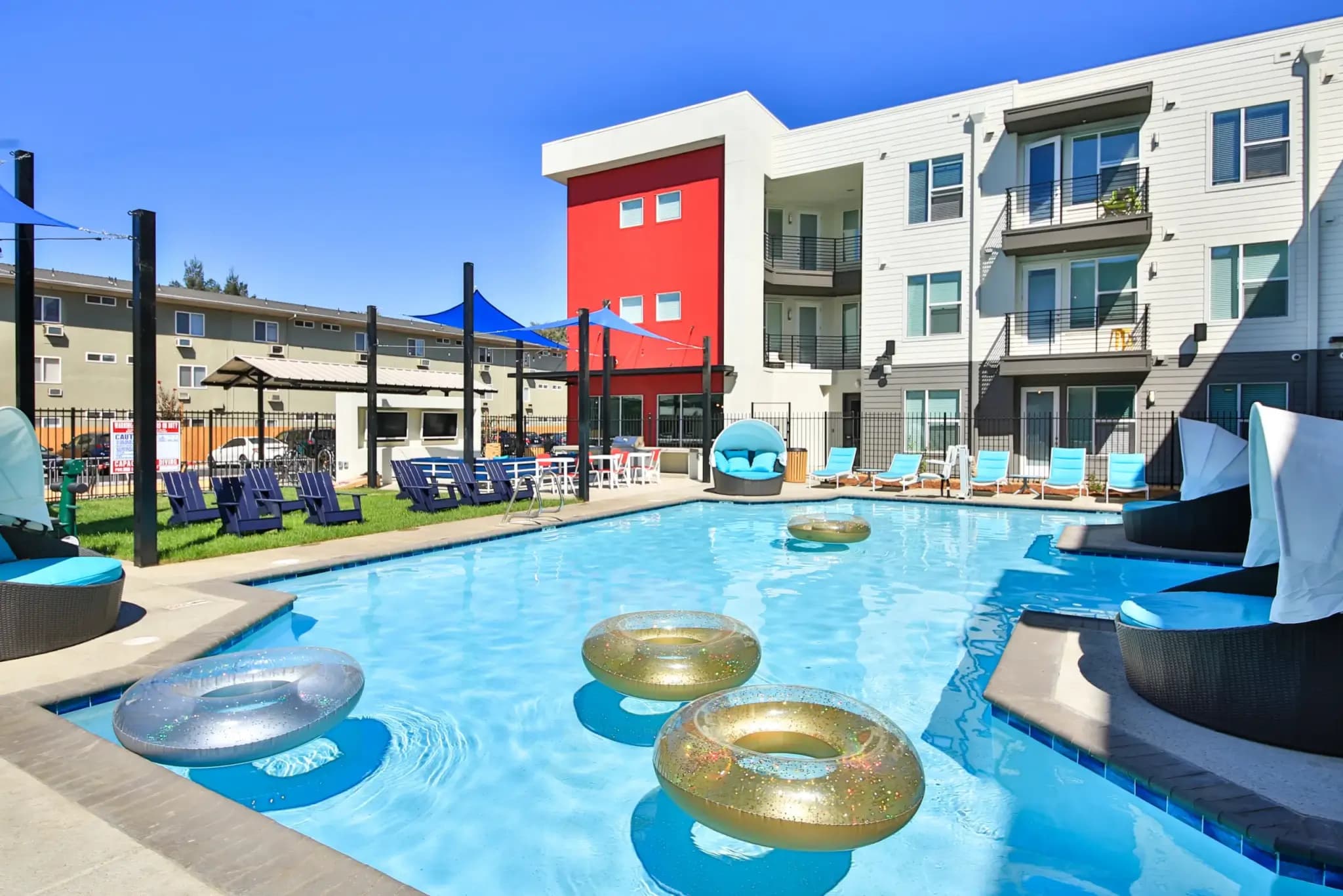 A modern outdoor swimming pool with floaties is surrounded by lounge chairs, cabanas, and shaded seating areas at Campus Walk Chico. A three-story apartment building with balconies overlooks the pool under a clear blue sky.