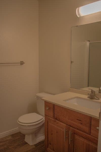 A simple bathroom in Carolina Townhomes Logan features a toilet, wooden vanity with sink, large mirror, and wall-mounted light. Light-colored walls and wood-like flooring complete the inviting space.