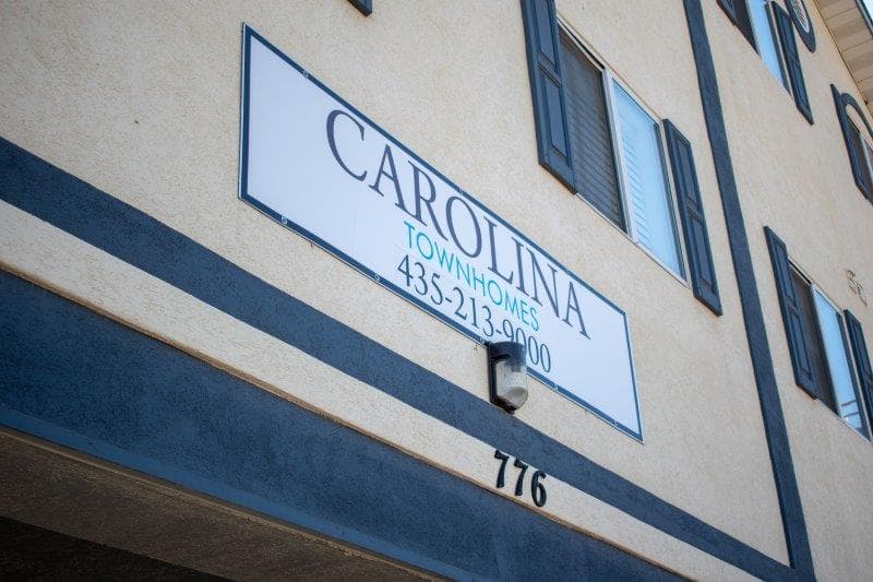 A sign on a beige building reads CAROLINA TOWNHOMES LOGAN 435-213-0000 above the address number 776. Black window shutters and several windows are visible in the background.