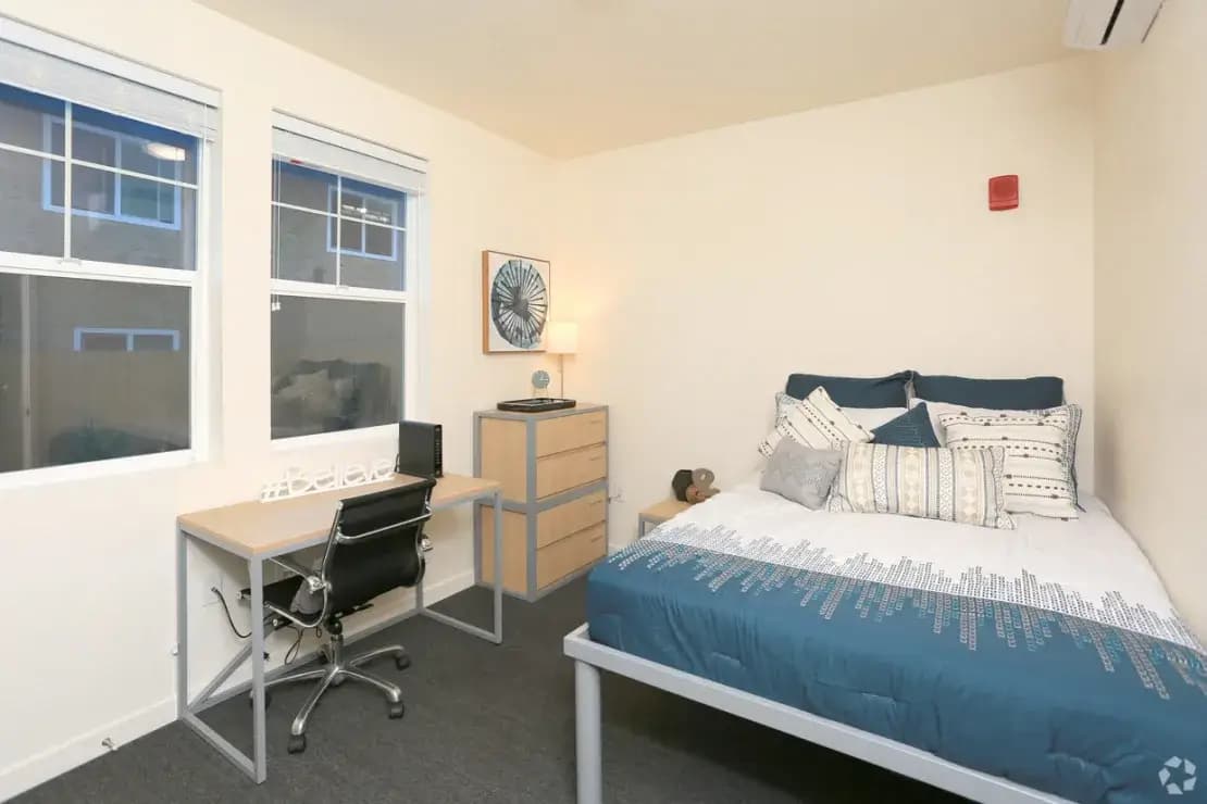 A tidy Cedar Flats Chico bedroom with a bed covered in blue and white bedding, a small wooden dresser with a lamp and decor, a desk with a chair and computer, and two windows letting in natural light.