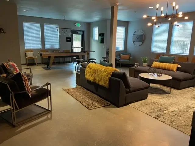 A modern lounge at Cedar Flats Chico features dark sofas, colorful pillows, a shaggy yellow throw, armchairs, a foosball table, round coffee tables, and large windows that flood the space with natural light. Contemporary lighting fixtures hang from the ceiling.
