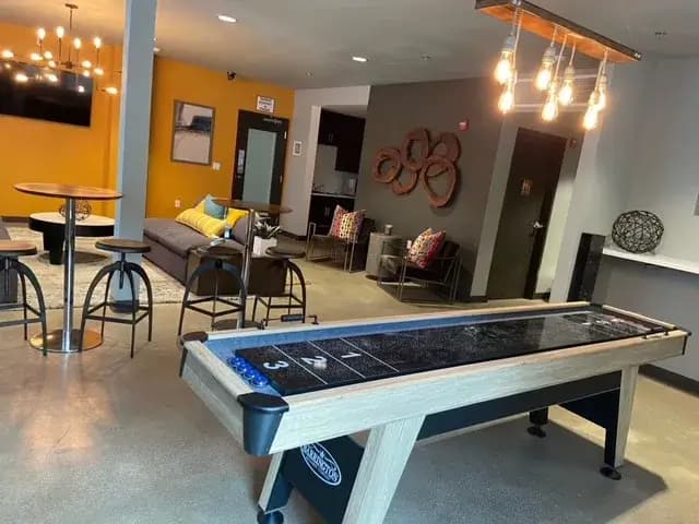 Modern lounge at Cedar Flats Chico featuring a central shuffleboard table, bar-style tables and stools, a sofa with yellow pillows, wall art, and decorative lighting. Walls are painted in stylish gray and yellow tones.