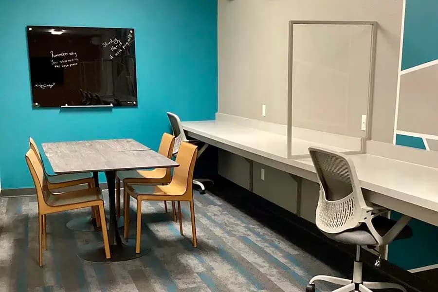 A modern study room at Cedar Flats Chico features a table with four chairs, a wall-mounted black glass board with writing, and a long desk with office chairs and a clear divider against a teal and gray wall.
