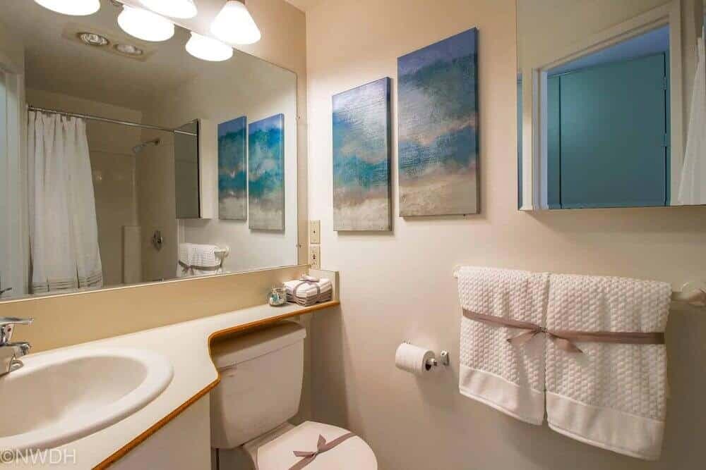 A small bathroom with a sink, toilet, and tub at The Collegian Eugene. Above the toilet are three ocean-themed paintings. Two textured white towels with ribbons hang on the wall, and a mirror reflects the room’s details.