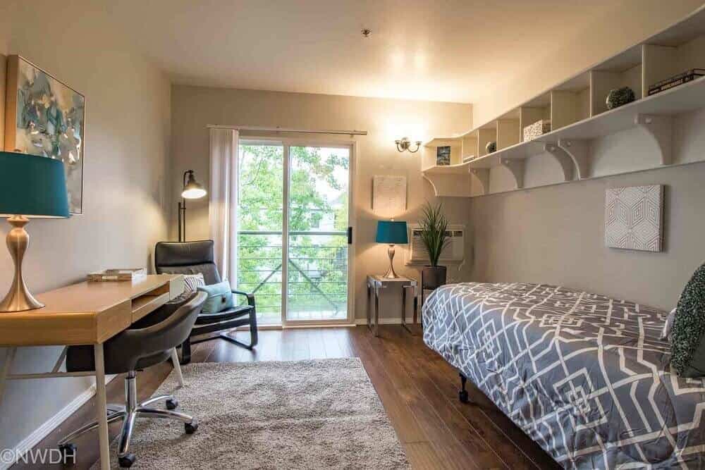 A bright, modern dorm room at The Collegian Eugene features a single bed, desk and chair, armchair by the window, wall shelves above the bed, a lamp, and a sliding glass door opening to a view of trees outside.