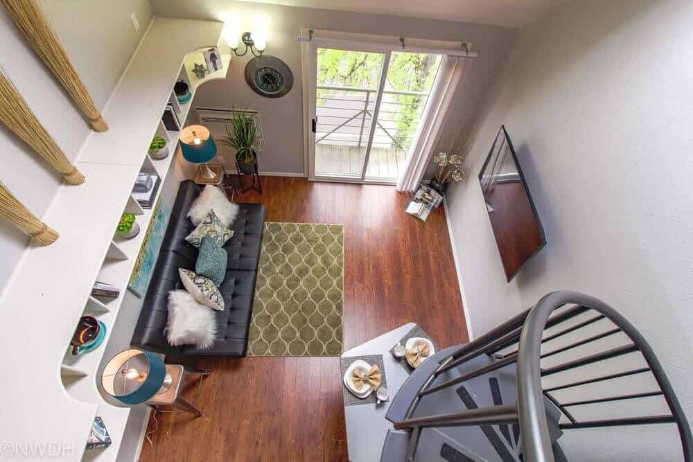 A modern loft living room at The Collegian Eugene features wood floors, a black futon with pillows, two blue lamps, a green rug, wall-mounted TV, bookshelf, and spiral staircase. Sliding glass doors open to a balcony.