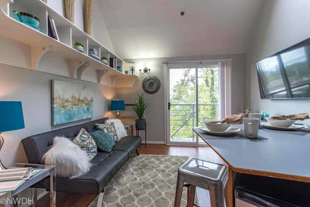 A modern, compact living room at The Collegian Eugene features a black futon with blue and white pillows, a geometric rug, wall art, open shelving, and a TV. Sunlight pours in through glass doors leading to a balcony with tree views.