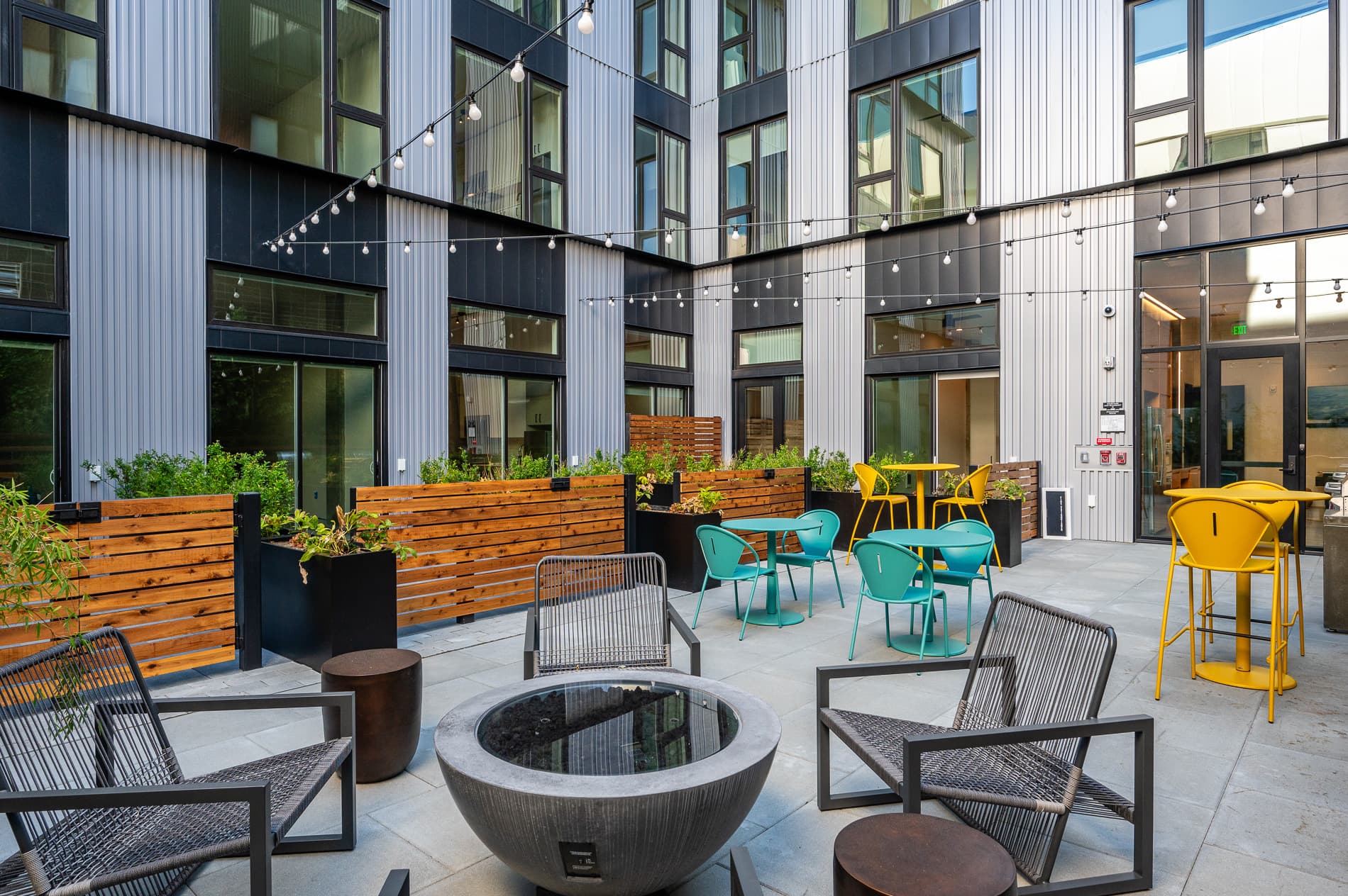 Modern outdoor patio at MERX Slabtown with string lights, metal chairs around a fire pit, colorful yellow and teal tables and chairs, wooden planters with greenery, and large windows on a multi-story building.