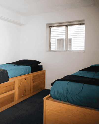 A small bedroom at Davenport Rexburg with two wooden twin beds, each with teal bedding and black pillows, placed against opposite walls under a window with blinds. White walls and a dark carpet complete the cozy look.