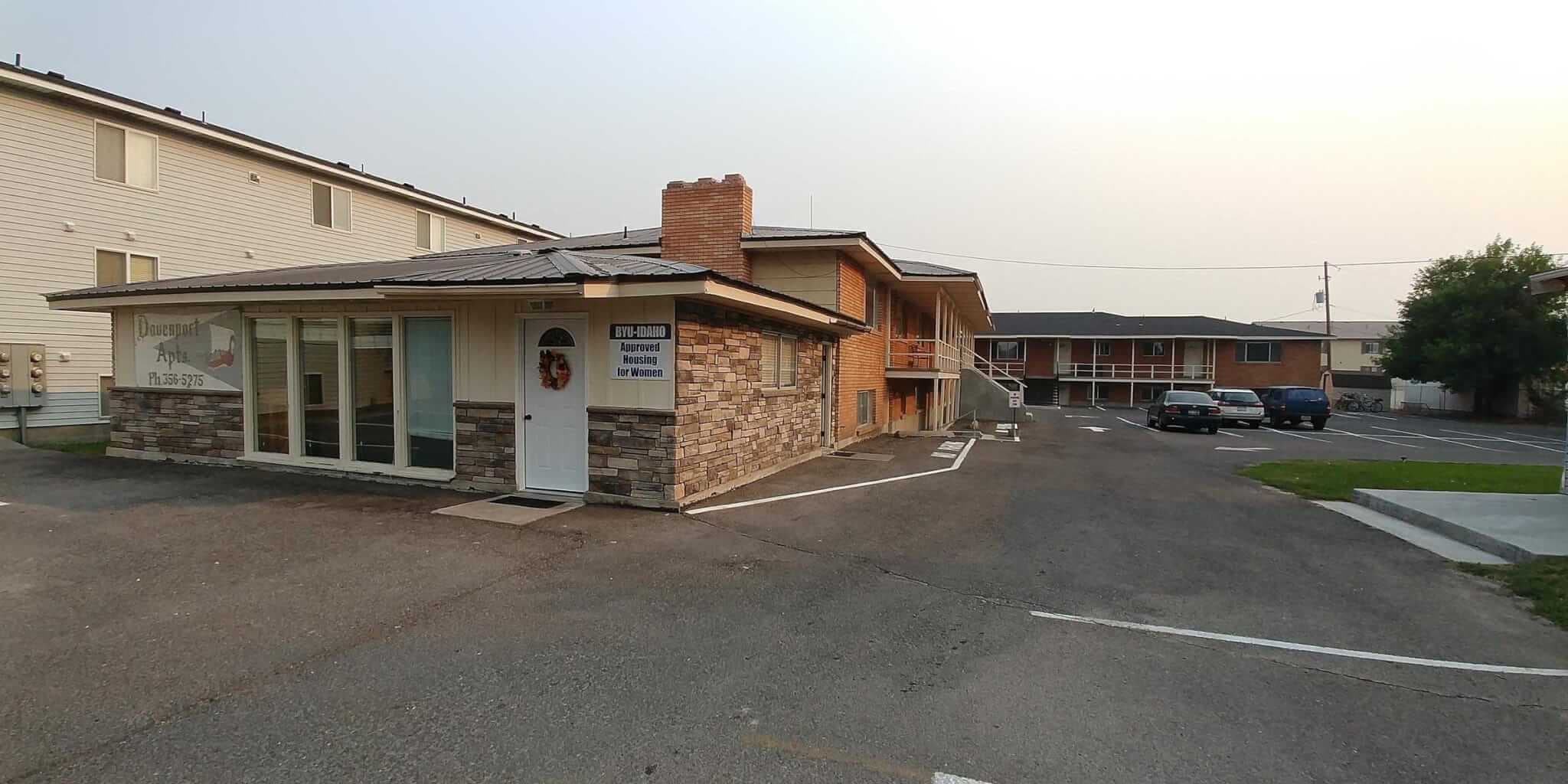 A small brick and stone office building with a sign reading Management Office, next to the Davenport Rexburg two-story apartment complex with parked cars, on a paved lot under a hazy sky.