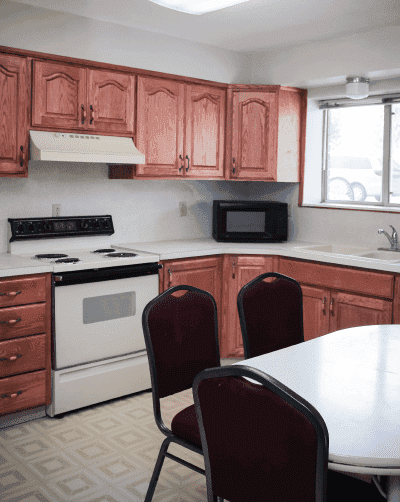 A small Davenport Rexburg kitchen features wooden cabinets, a white stove, range hood, microwave, and a sink beneath a window. In the foreground, a white table is surrounded by four maroon chairs.