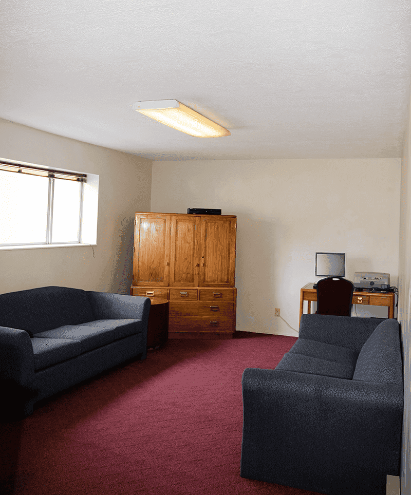 A sparsely furnished living room in Davenport Rexburg features two dark sofas, a wooden cabinet, a desk with a computer and printer, and a window letting in natural light on the left. The carpet is red and the walls are light-colored.
