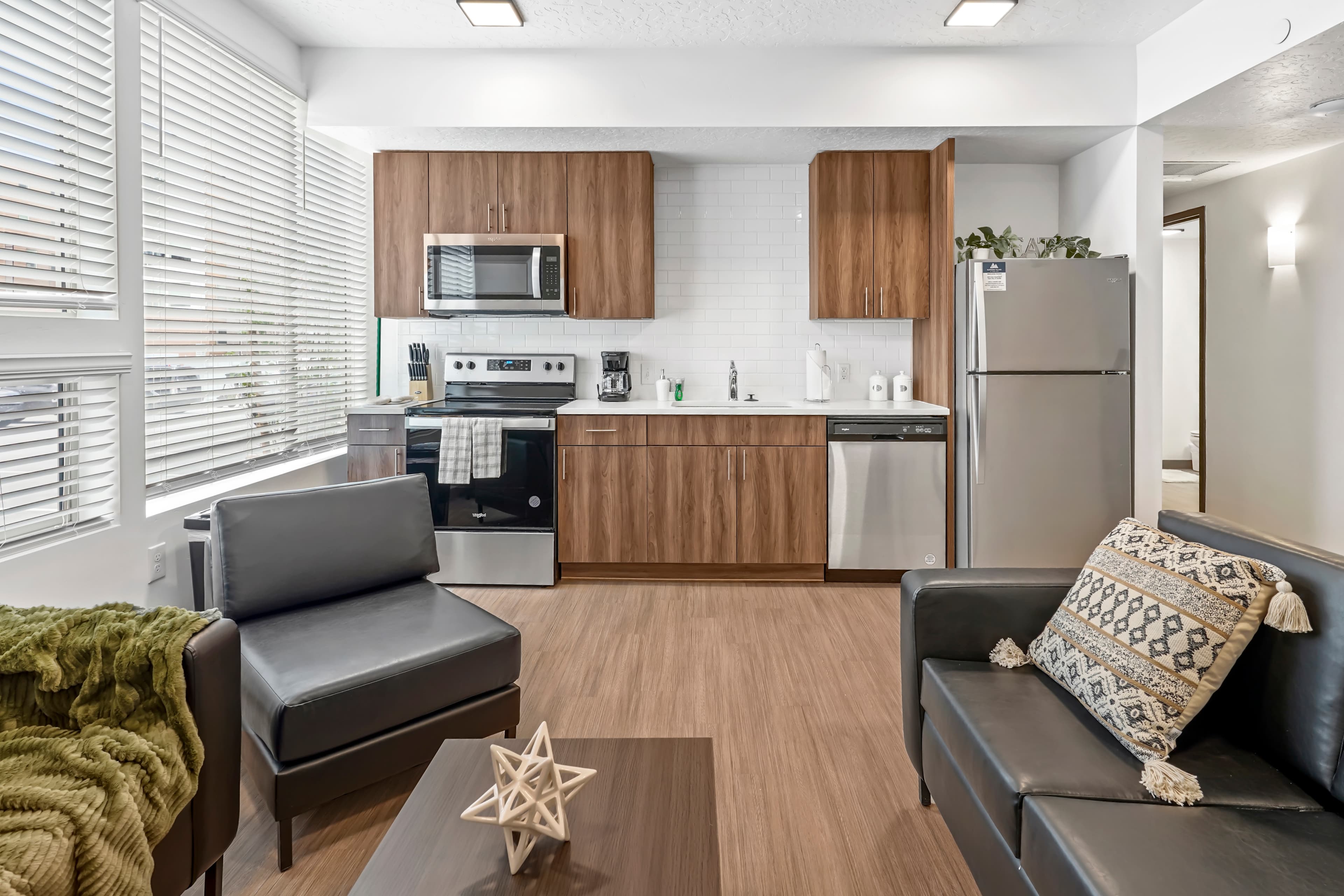 Modern apartment living room and kitchen in Slopeside Village Park City, featuring wood cabinets, stainless steel appliances, black leather sofas, a patterned pillow, a coffee table with decor, and large windows that fill the space with natural light.