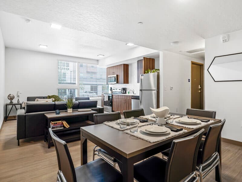 Modern open-concept apartment at Slopeside Village Park City featuring a dining table set for six, a cozy living area with couch and TV stand, and a kitchen with stainless steel appliances. Large windows fill the space with natural light.