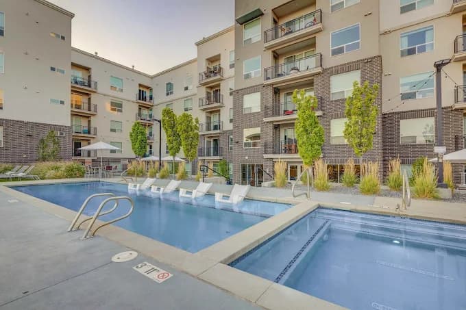 Outdoor pool area at a modern apartment complex, featuring lounge chairs partially submerged in the shallow end, surrounded by multi-story buildings with balconies and landscaping.