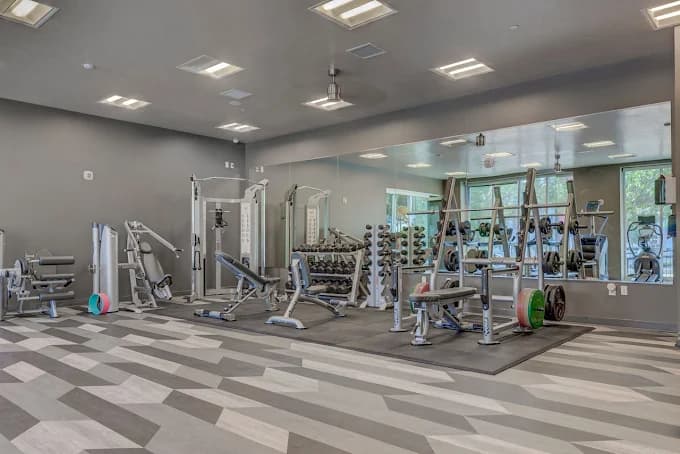 Modern gym with weight benches, free weights, resistance machines, and a mirrored wall. The room has geometric patterned flooring and large windows letting in natural light.