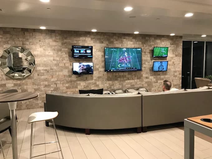 A man sits on a curved gray couch in a modern lounge, watching multiple TVs on a brick wall, including a large screen showing a football game and four smaller screens with various programs.