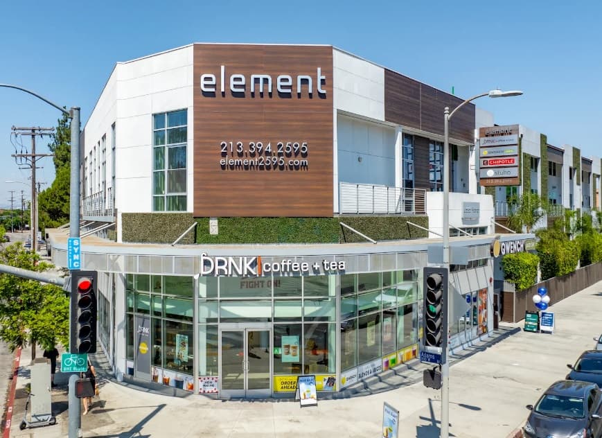 A modern, two-story building with “Element by Stuho” signage on the upper facade, featuring large windows and housing DRNK coffee + tea on the ground floor. Traffic lights and street signs are visible at the intersection outside.