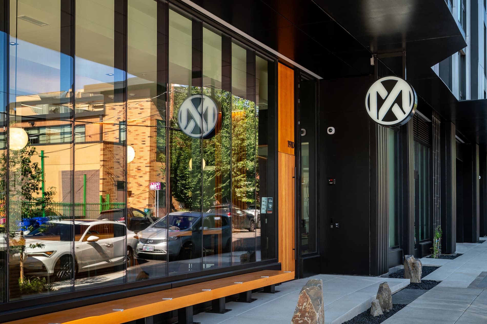 A modern building with large glass windows reflects cars and nearby structures in Slabtown. A round MERX sign with overlapping “M” and “A” letters hangs above a wooden bench along the sidewalk.