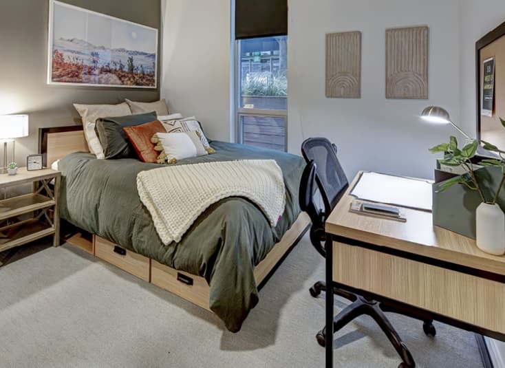 A modern bedroom in the Theory U District features a neatly made bed with green bedding and a cream knit blanket. A wooden desk with a chair, lamp, and plant sits nearby, while wall art above the bed and a window brighten the space.