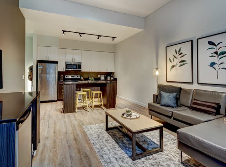 Modern open-concept living room and kitchen with stainless steel appliances, white cabinets, a dark island with yellow stools, gray sectional sofa, decorative pillows, coffee table, and botanical wall art.