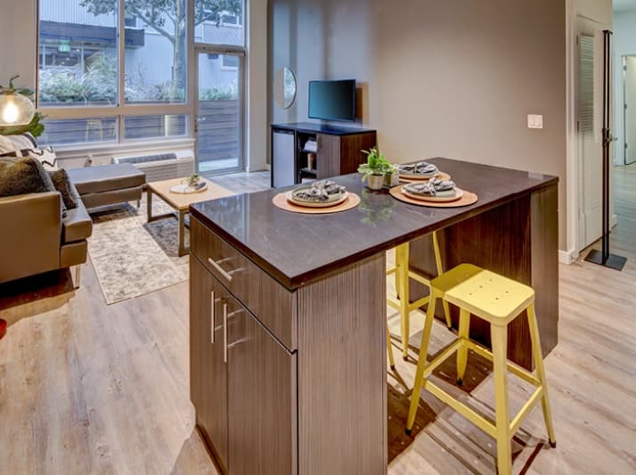 Kitchen island with dark countertop and two yellow stools set for dining, open to a living area with a couch, coffee table, TV, and large window letting in natural light. Light wood floors throughout the modern space.