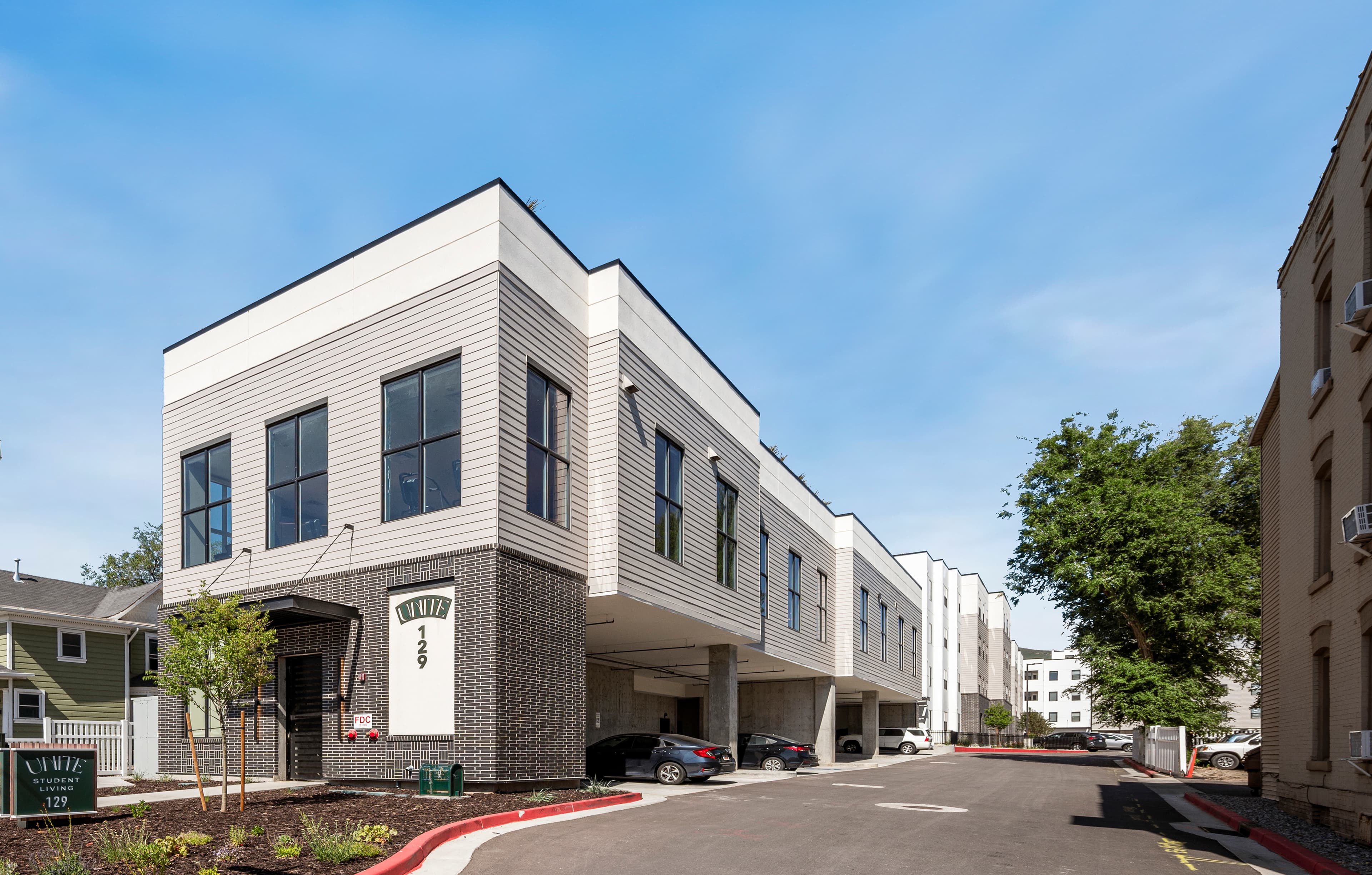 Modern apartment building with a white and gray exterior, large windows, and covered parking spaces underneath—ideal for Unite Student Housing Salt Lake City. Several cars are parked beside the address “129” sign and landscaped greenery under a blue sky.