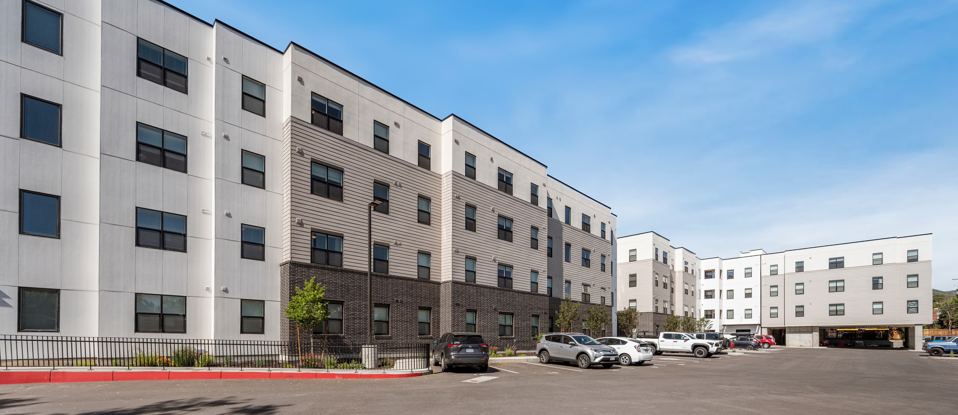 A modern apartment complex, ideal for Unite Student Housing Salt Lake City, with three- and four-story buildings, white and gray exteriors, black-framed windows, and a parking lot full of cars beneath a clear blue sky.