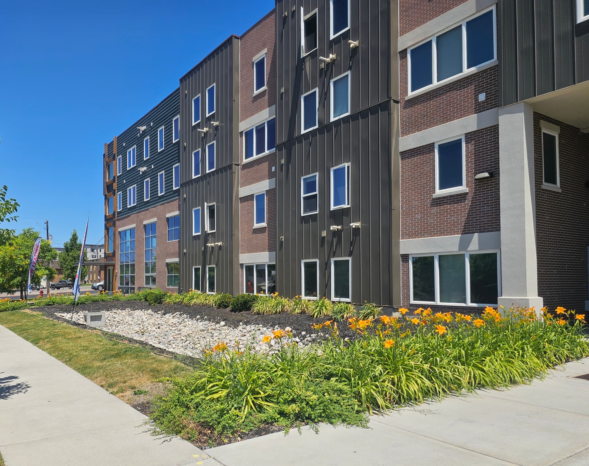 A modern, multi-story apartment building—The Factory Student Housing Logan—features brown and black exterior panels, large windows, and a landscaped area with orange flowers and greenery along the sidewalk under a clear blue sky.