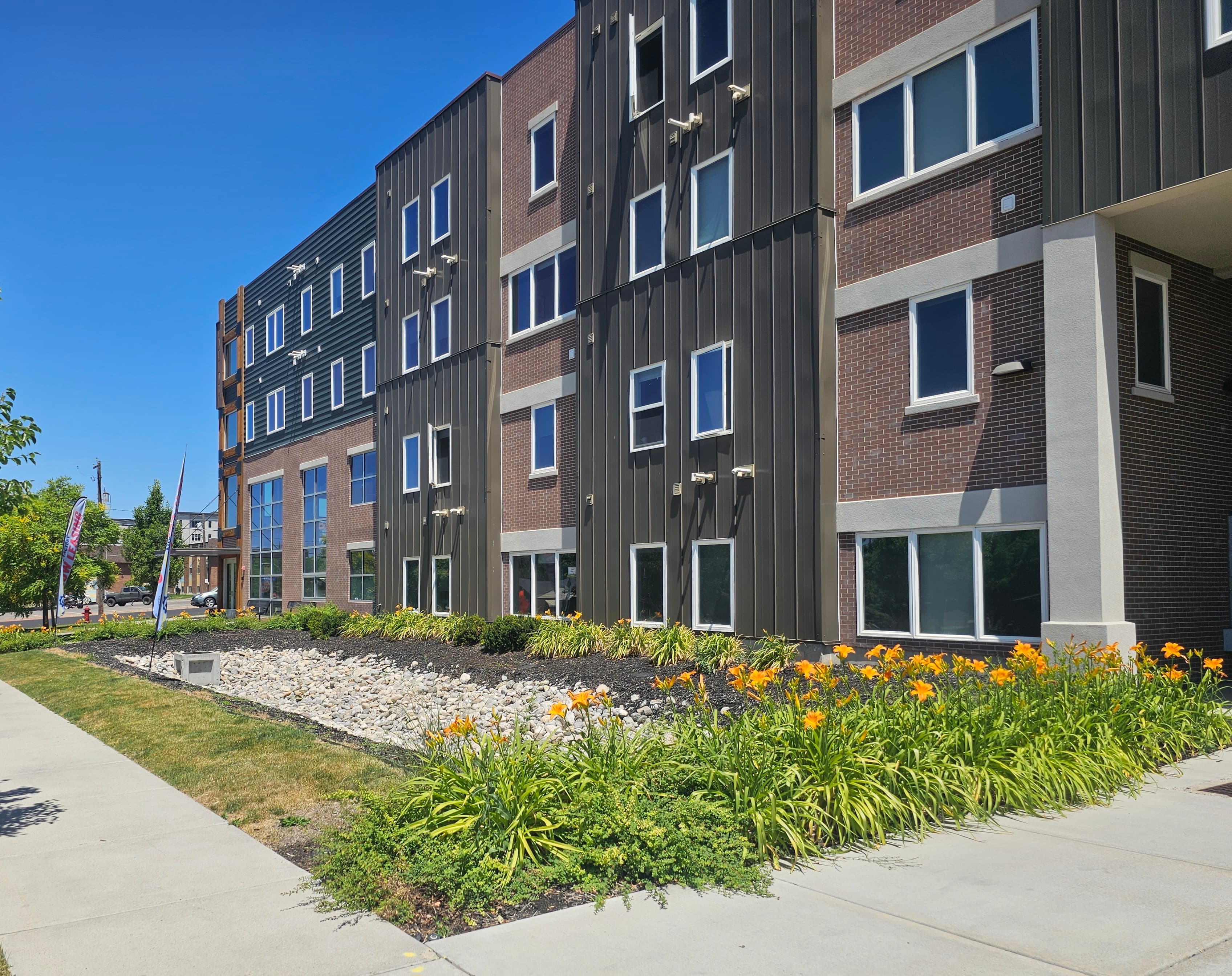 A modern, multi-story apartment building—The Factory Student Housing Logan—features brown and black exterior panels, large windows, and a landscaped area with orange flowers and greenery along the sidewalk under a clear blue sky.