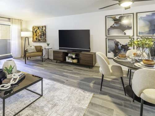 Modern living and dining room at Foothill Place Salt Lake City features gray wood flooring, a large TV on a dark console, light chairs around the dining table, abstract wall art, a standing lamp, and plenty of natural light from large windows.