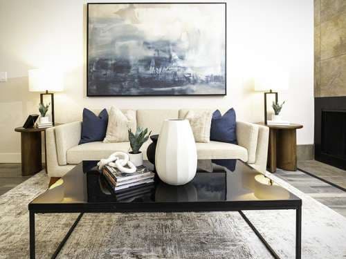 A modern living room at Foothill Place Salt Lake City features a cream sofa, blue and beige pillows, side tables with lamps, a large abstract painting, and a black coffee table adorned with decor items and books.