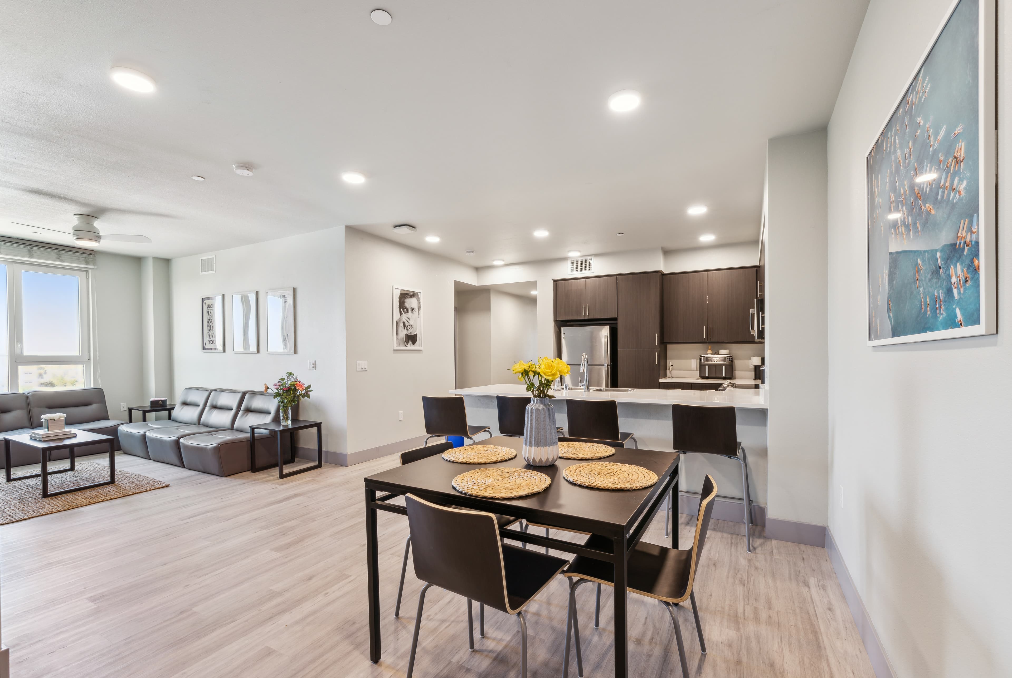 Modern open-concept apartment with a dining area set for four, contemporary kitchen with dark cabinets, and a cozy living room with a gray sofa. Neutral decor and bright natural light fill the space.